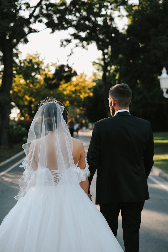 Newlywed couple walking hand in hand through a sunlit park, captured from behind.